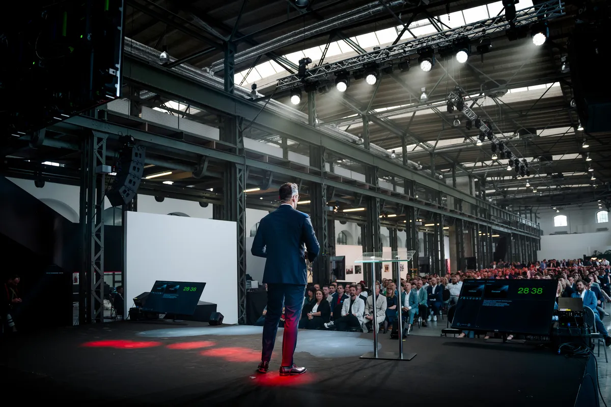 Řečník na pódiu ve velkém industriálním sále plném hostů